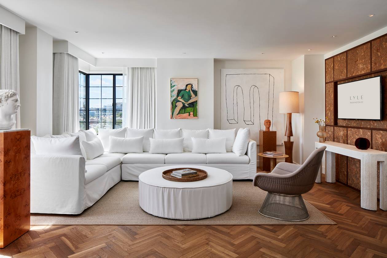 A bright living room in the Lyle Suite with a white sectional sofa, round ottoman, and modern artwork with neutral tones.