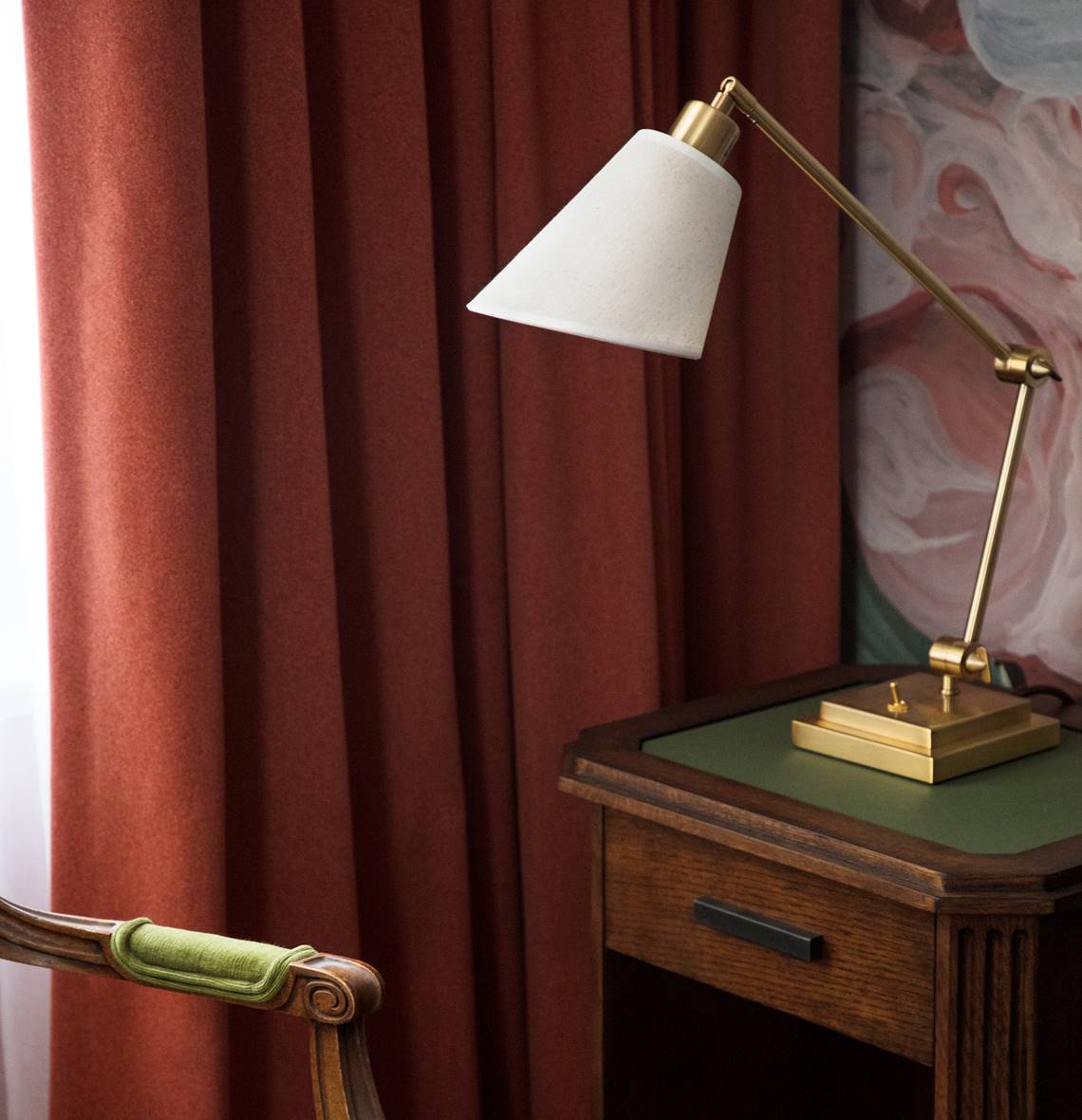 A classic wooden nightstand with a green top features a brass lamp with a white shade beside a deep red curtain.