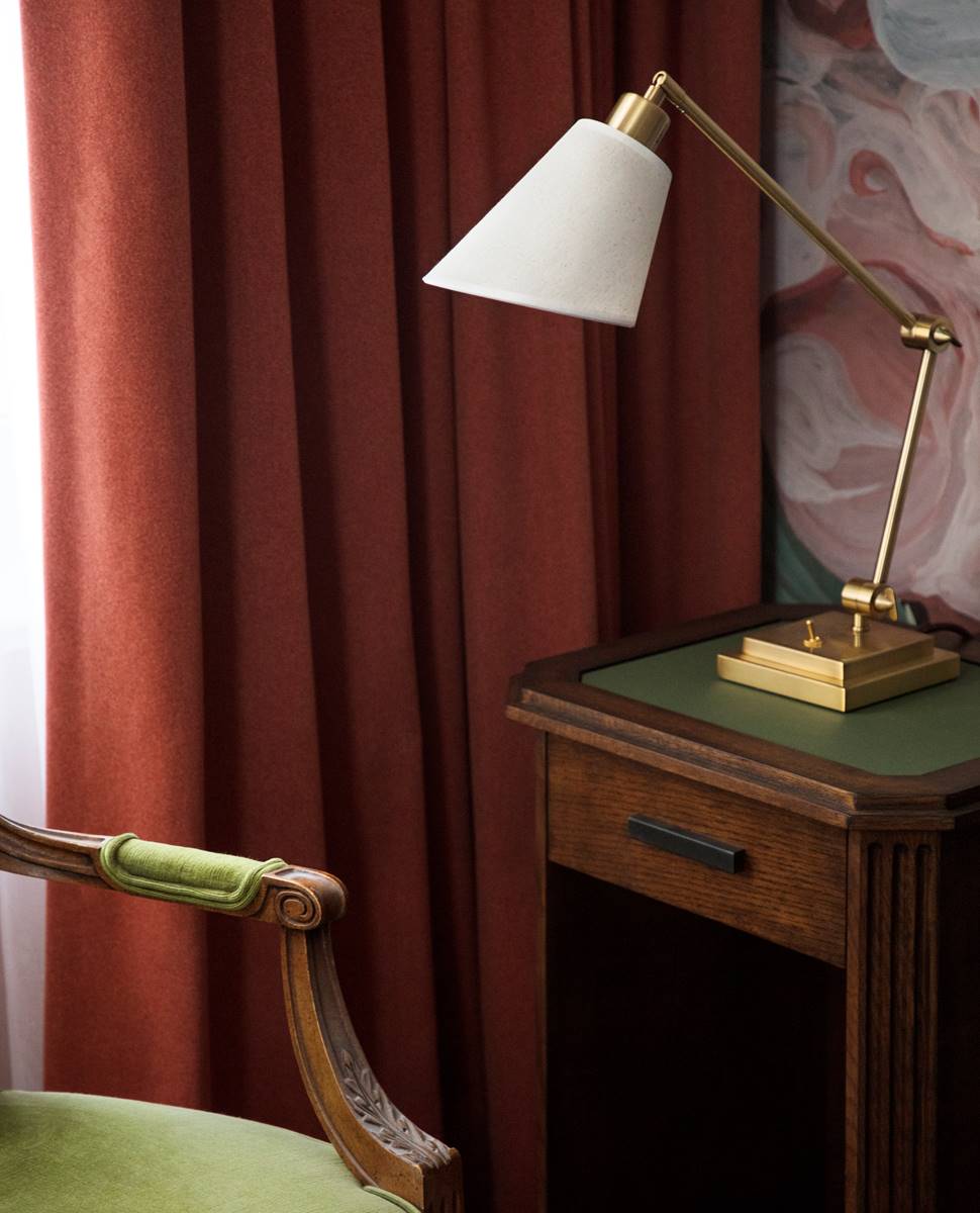 A classic wooden nightstand with a green top features a brass lamp with a white shade beside a deep red curtain.