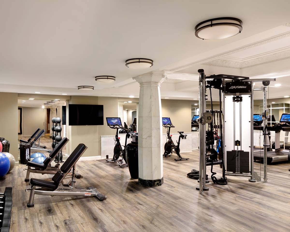 A well-equipped, modern gym with various exercise machines, free weights, and cardio equipment. Natural light fills the room.