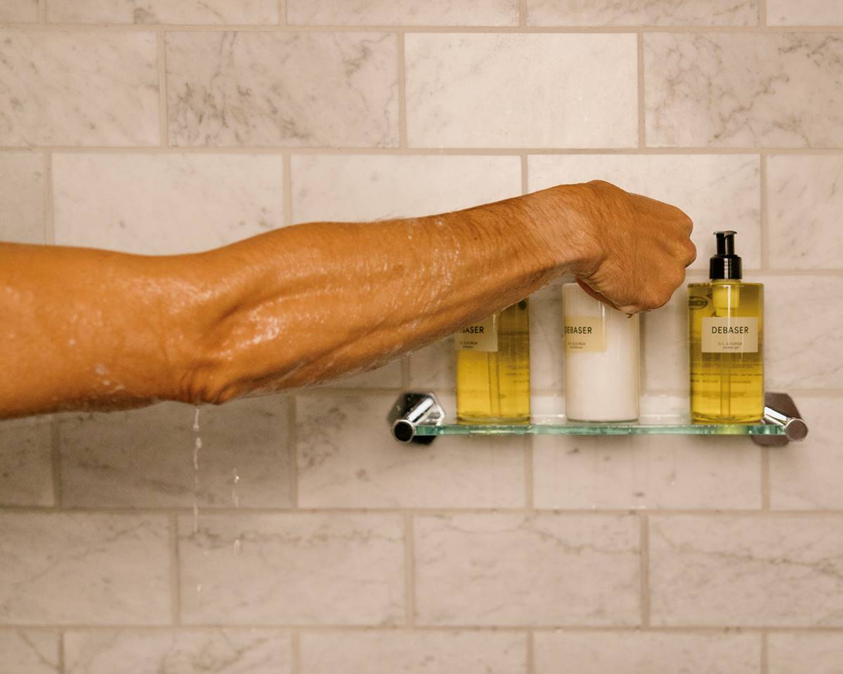 A wet arm reaches for a Debaser body care product on a glass shelf in a marble-tiled shower in a Riggs DC hotel bathroom.