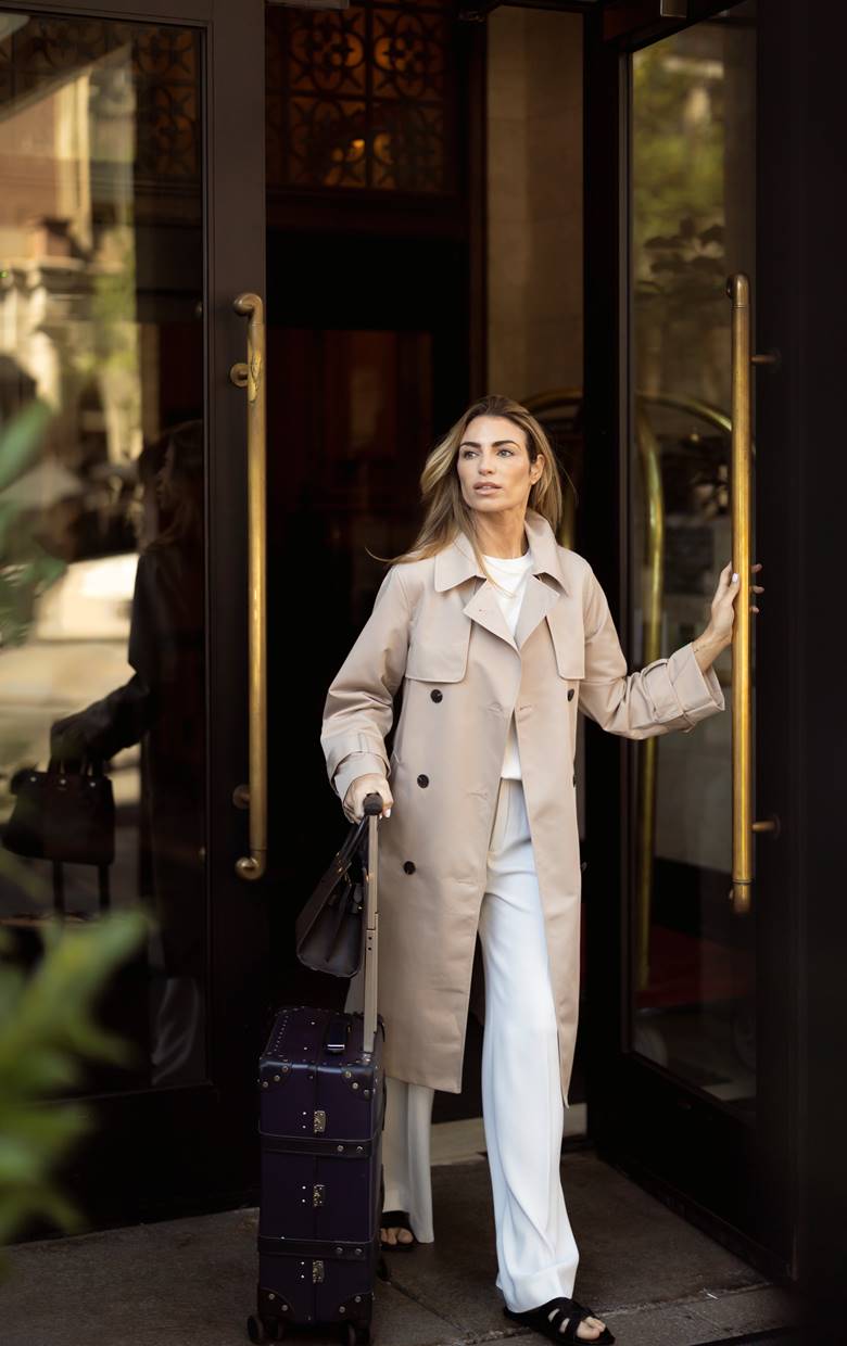 A woman in a beige trench coat and white outfit steps out of the Riggs DC hotel door, pulling a navy suitcase.