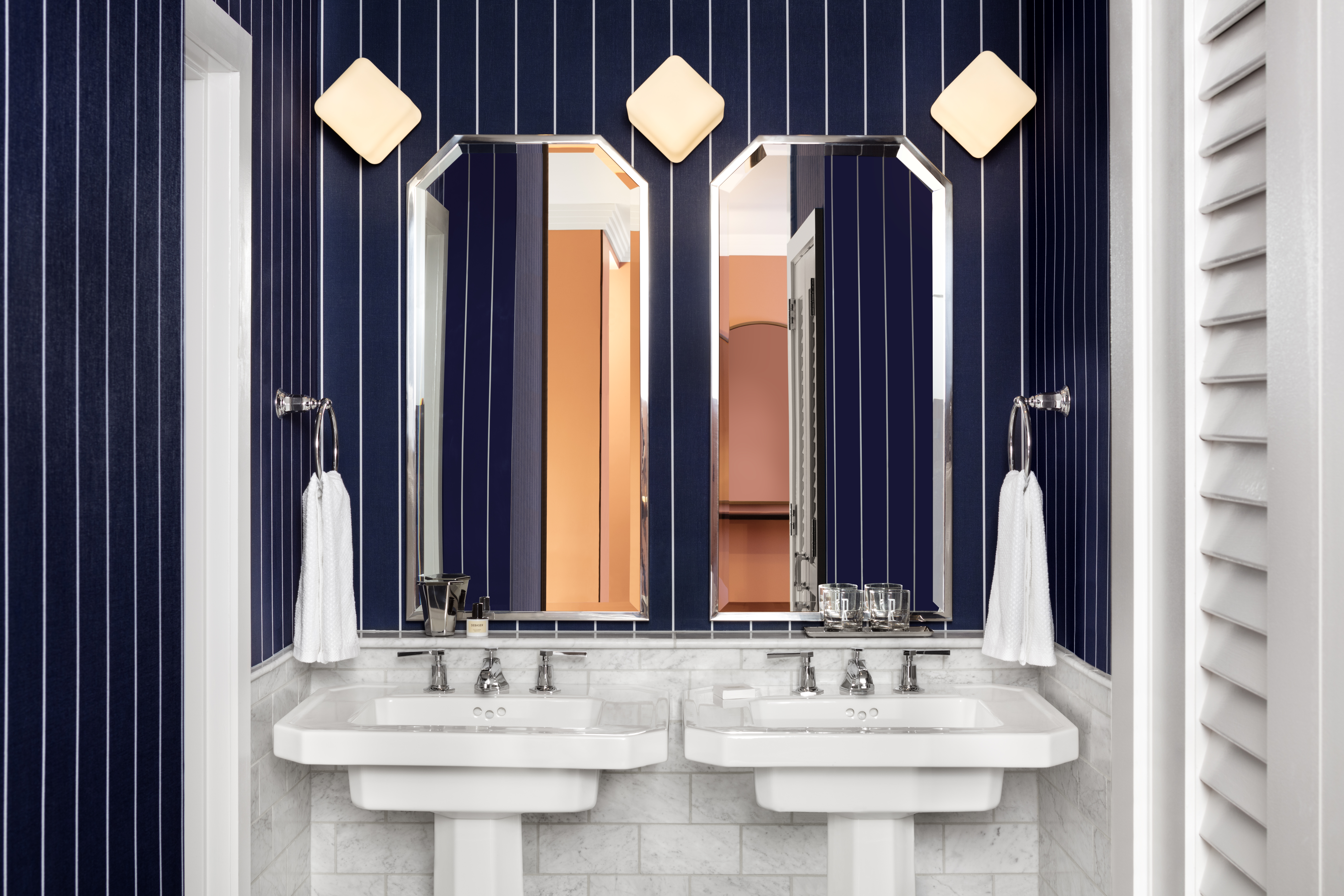 A bathroom with deep blue striped walls, twin pedestal sinks, and octagonal mirrors reflecting a warm-toned interior.