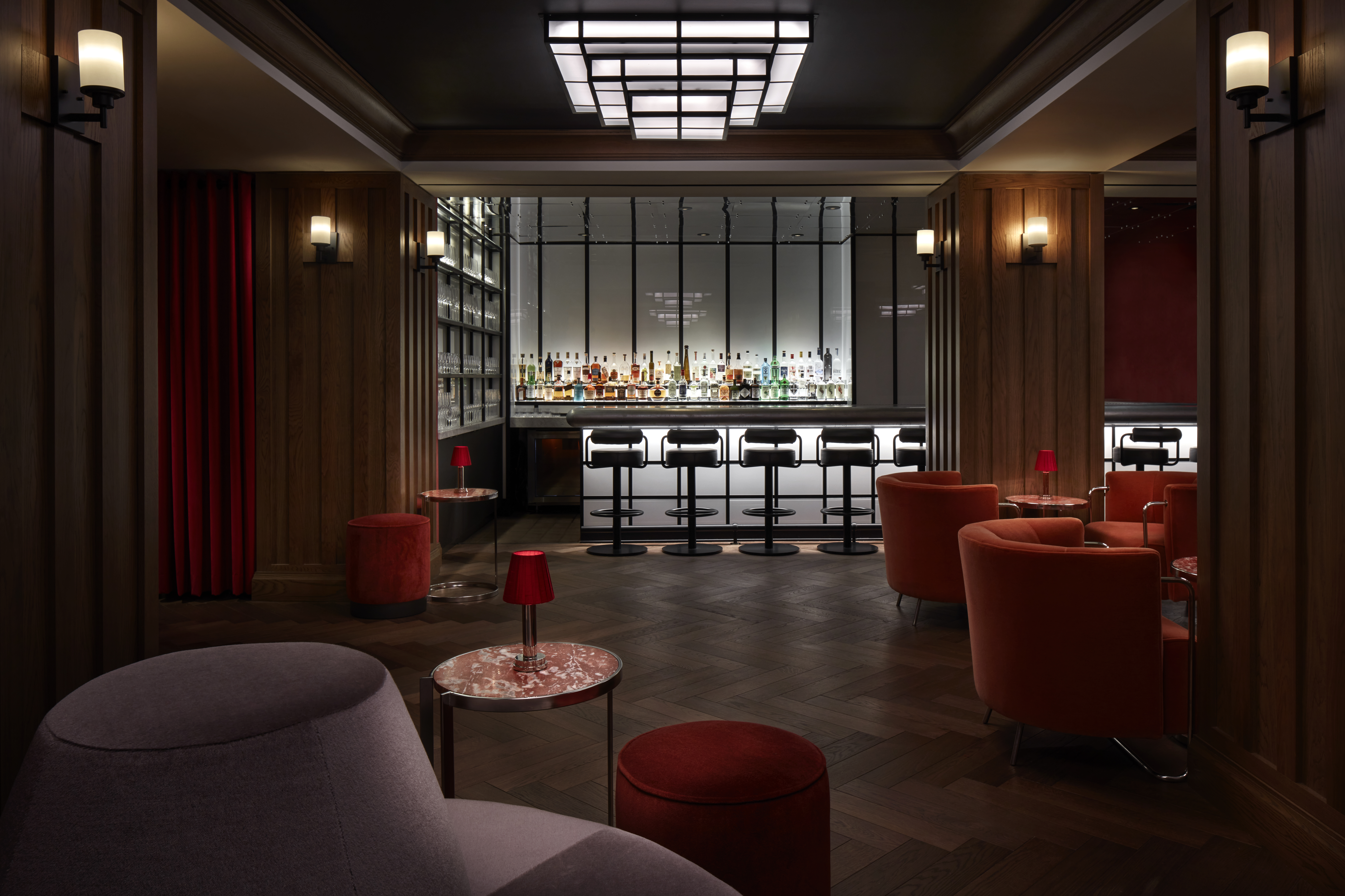 A dimly lit bar with a long counter and stools, liquor bottles on shelves, and modern decor.