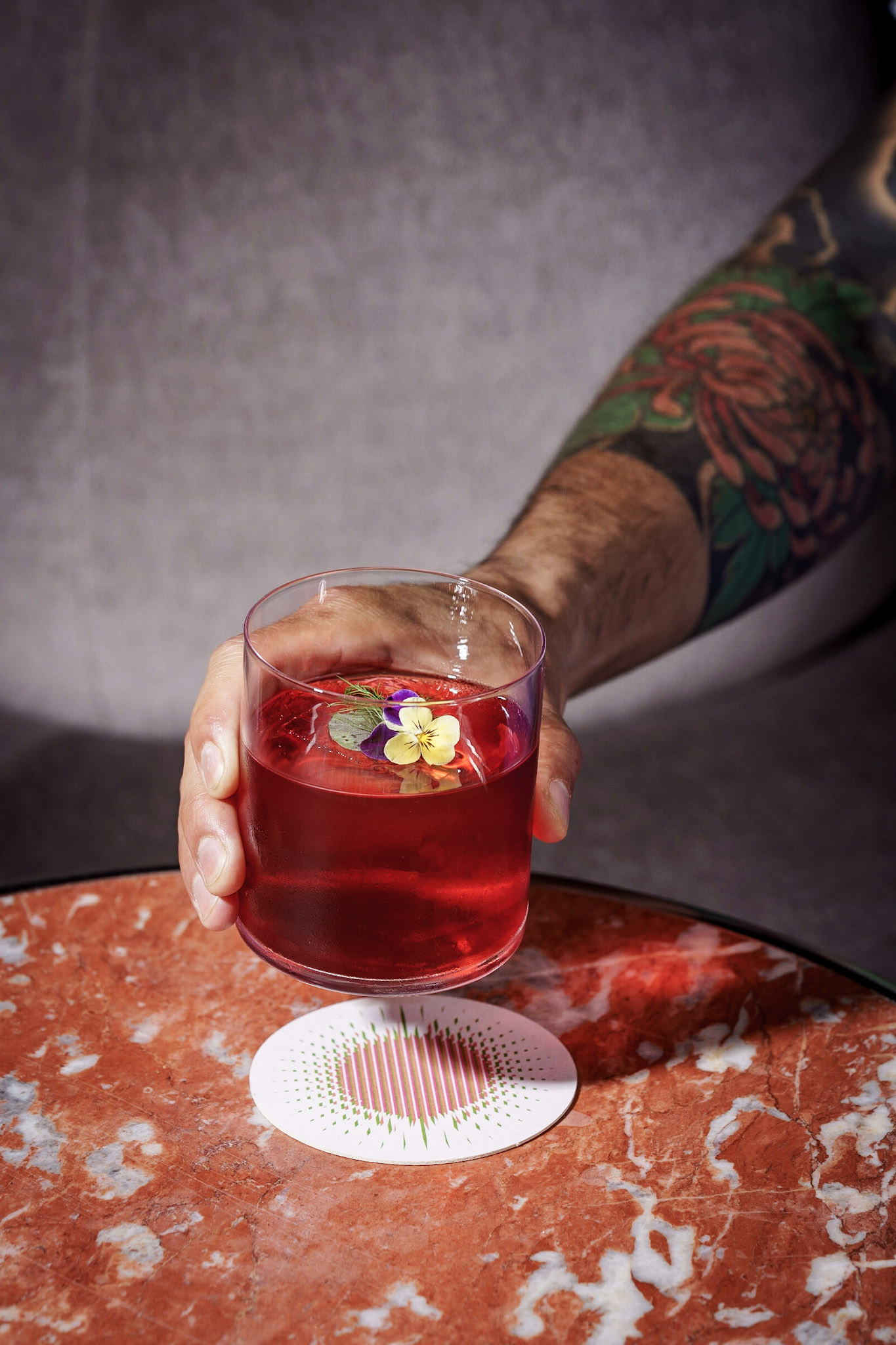 Hand holding a red cocktail garnished with flowers, set against a backdrop of a tattooed arm and a marble table.