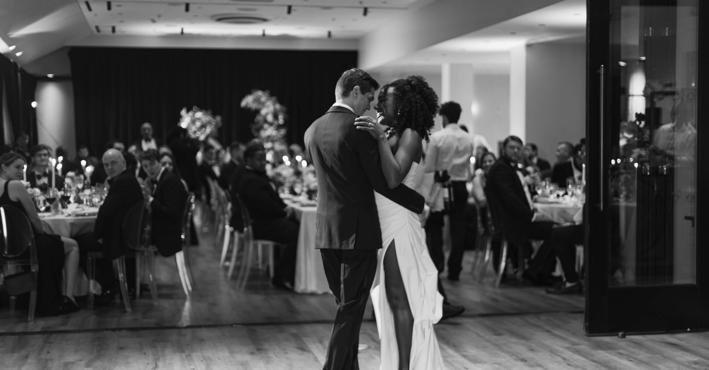 A black and white photo of a wedding reception with a couple dancing in the foreground and guests seated at tables in the background.