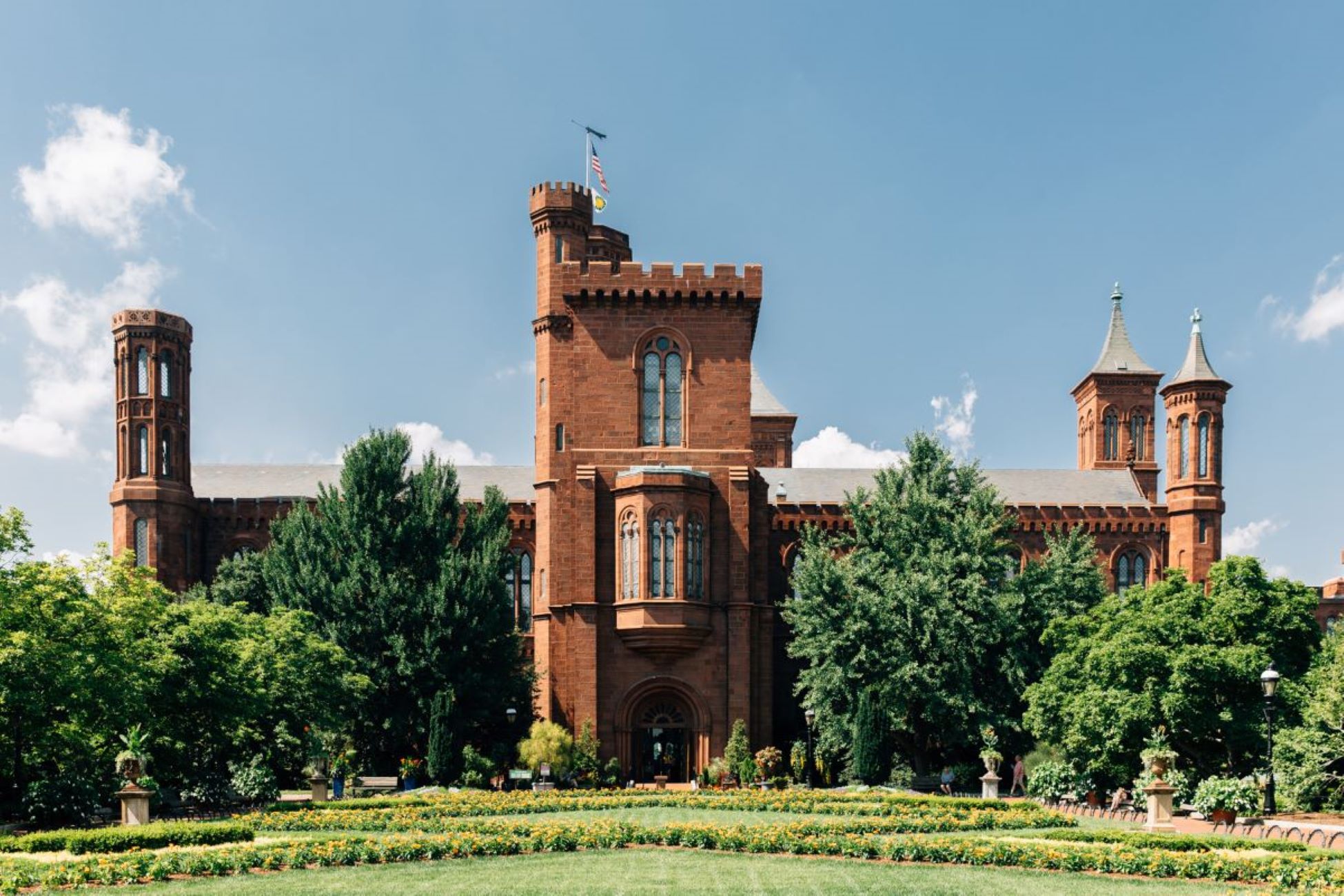 A historic red-brick castle with turrets and arched windows stands among lush gardens under a clear blue sky.