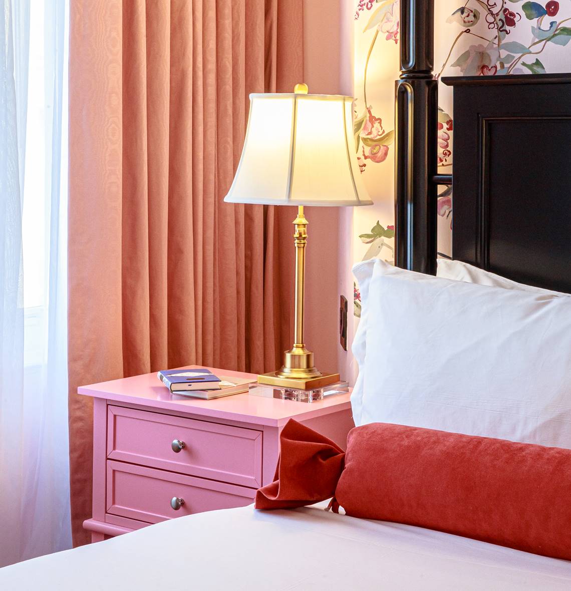 A pink-themed bedroom with a floral accent wall, a brass lamp, and a nightstand stacked with books.