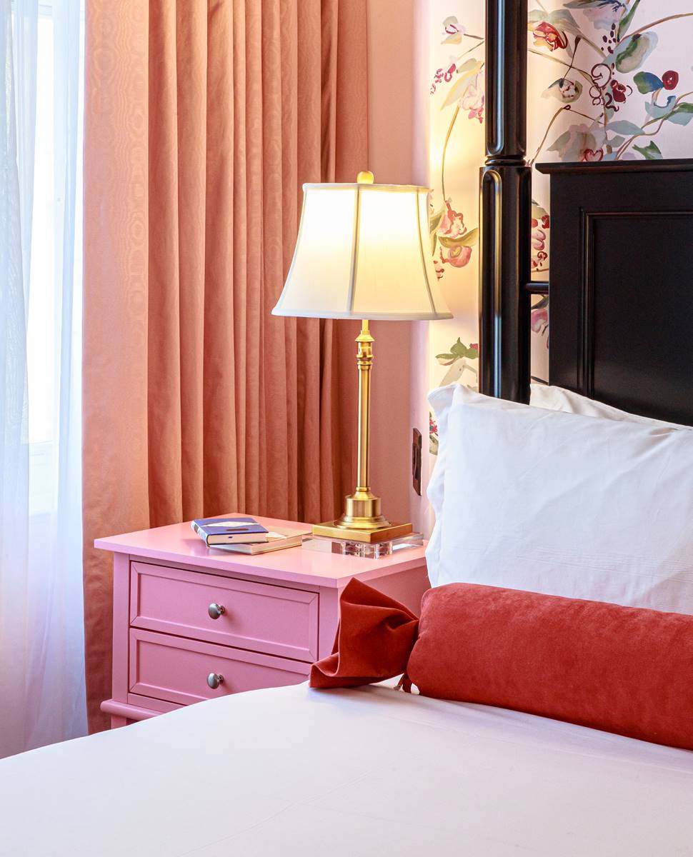A pink-themed bedroom with a floral accent wall, a brass lamp, and a nightstand stacked with books.