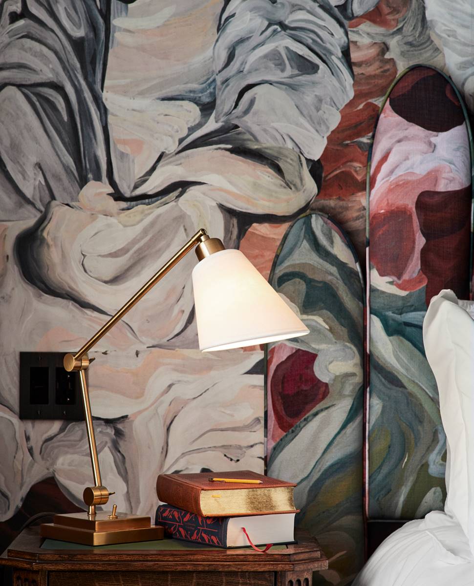 Bedside table at a Riggs DC hotel room with stacked books, a brass reading lamp, and abstract floral wallpaper backdrop.