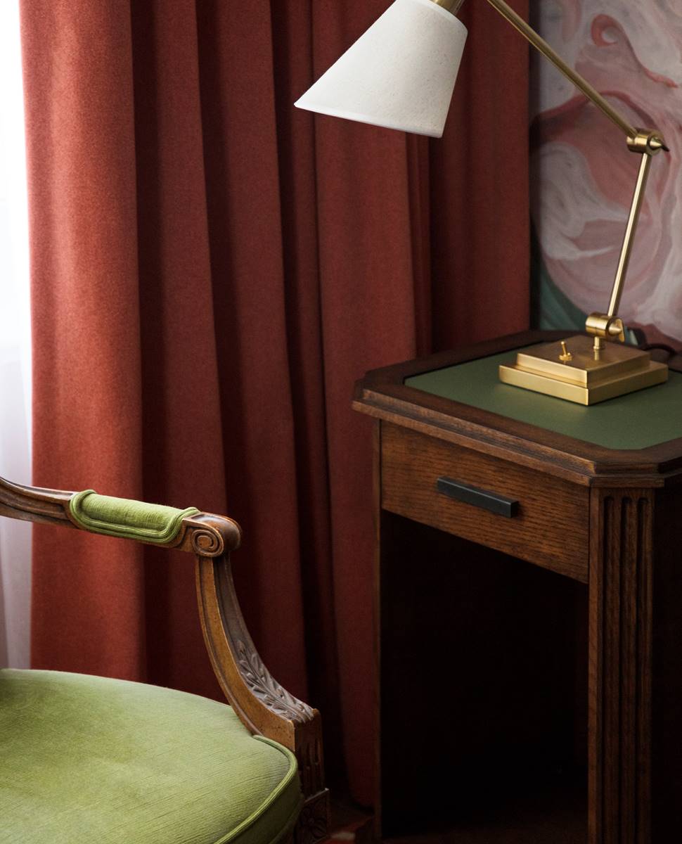 A classic wooden nightstand with a green top features a brass lamp with a white shade beside a deep red curtain.
