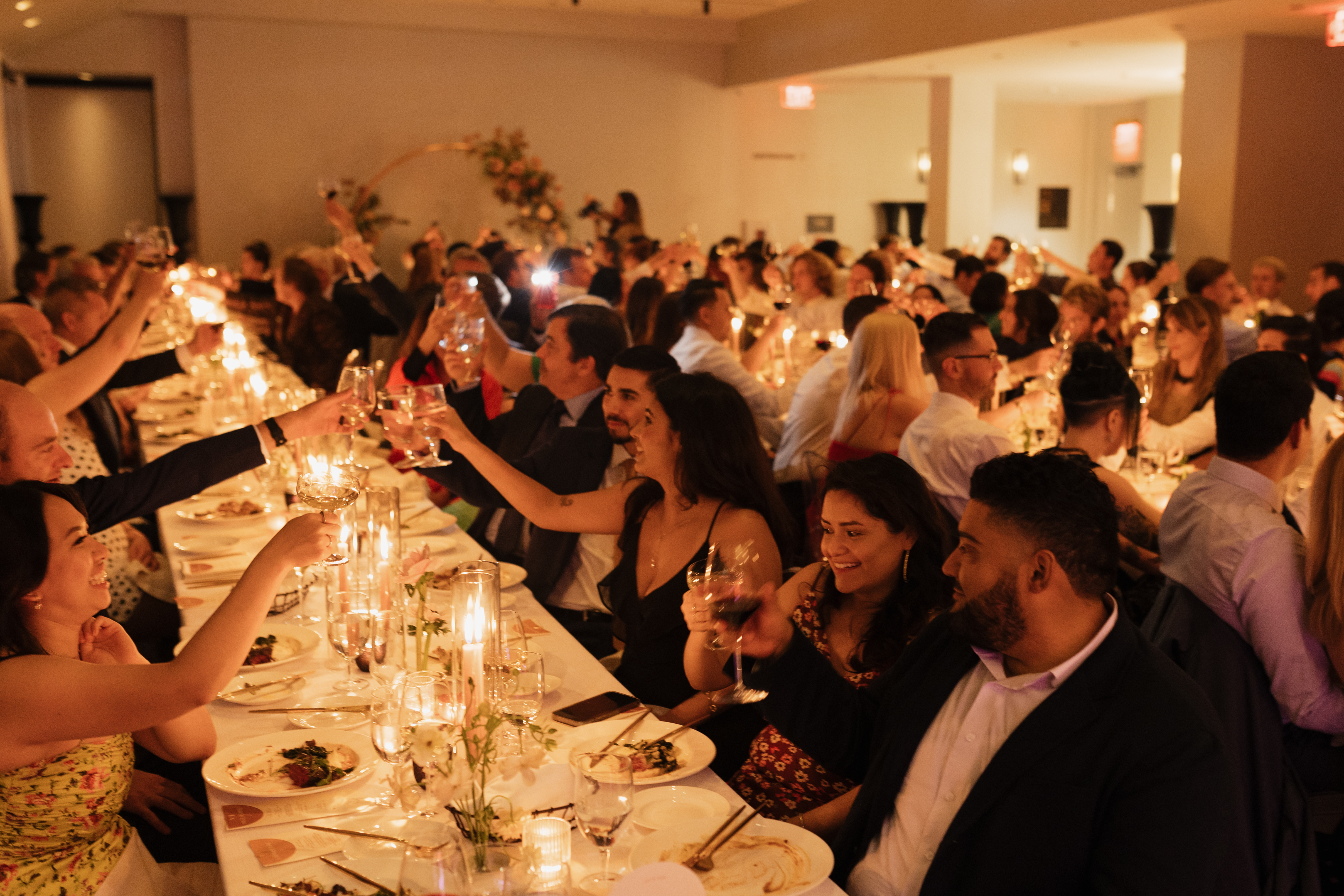 A large group of people sitting at a long table, raising their wine glasses in a toast.