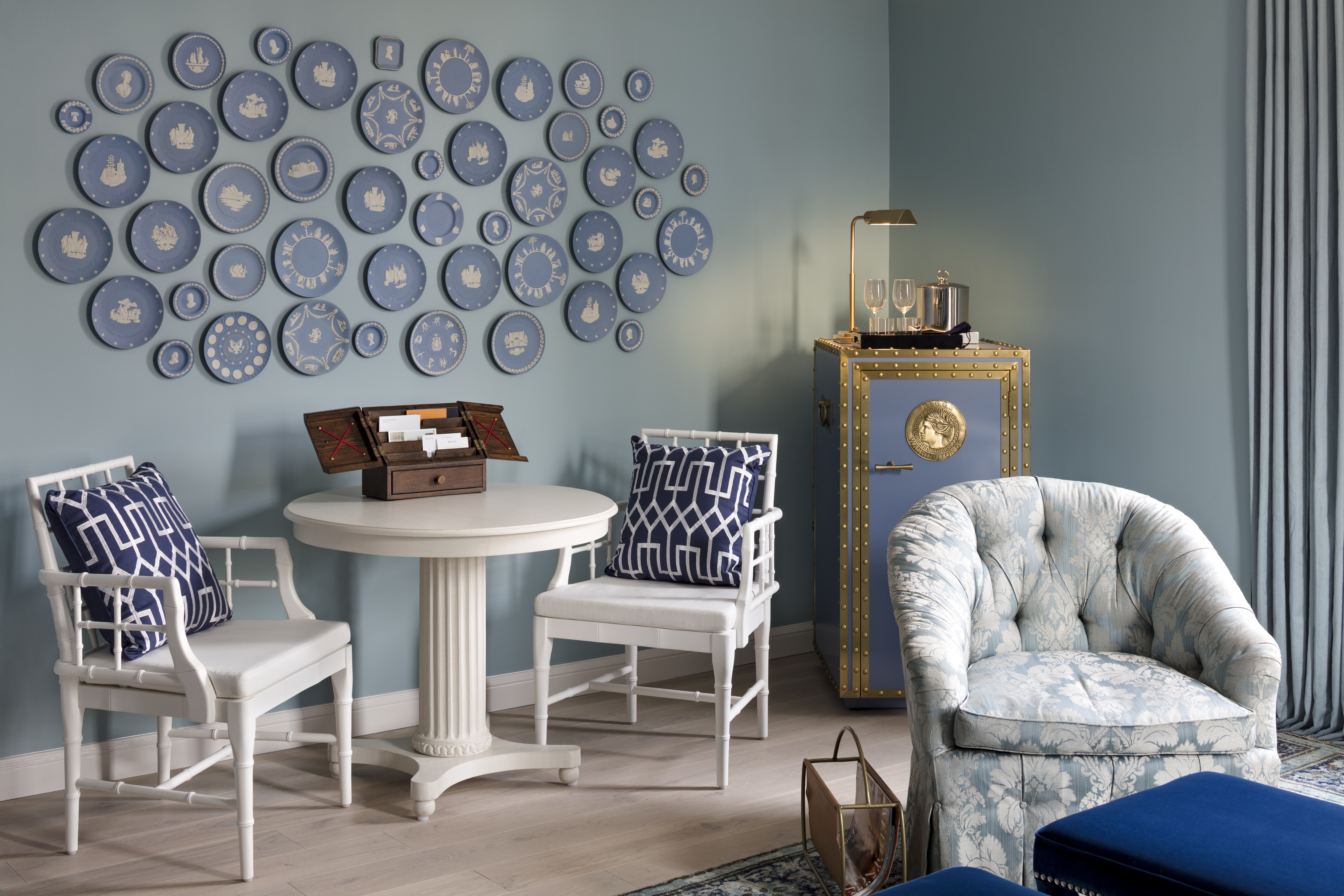 Seating area in a hotel room with blue-themed decor, including decorative plates on the wall, white chairs, and a safe.