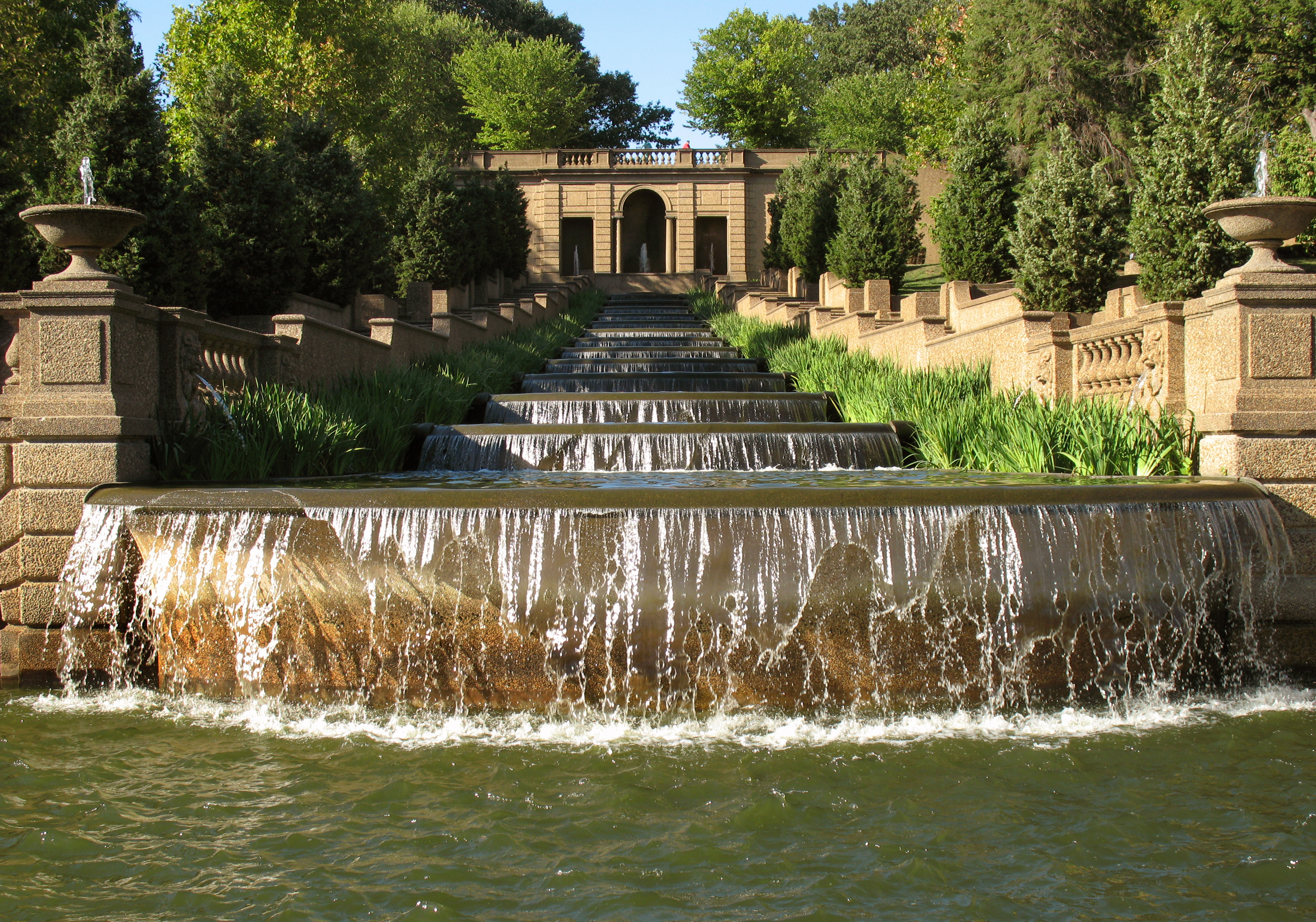 A cascading waterfall flows over a series of stone steps, surrounded by greenery and an elegant terrace.