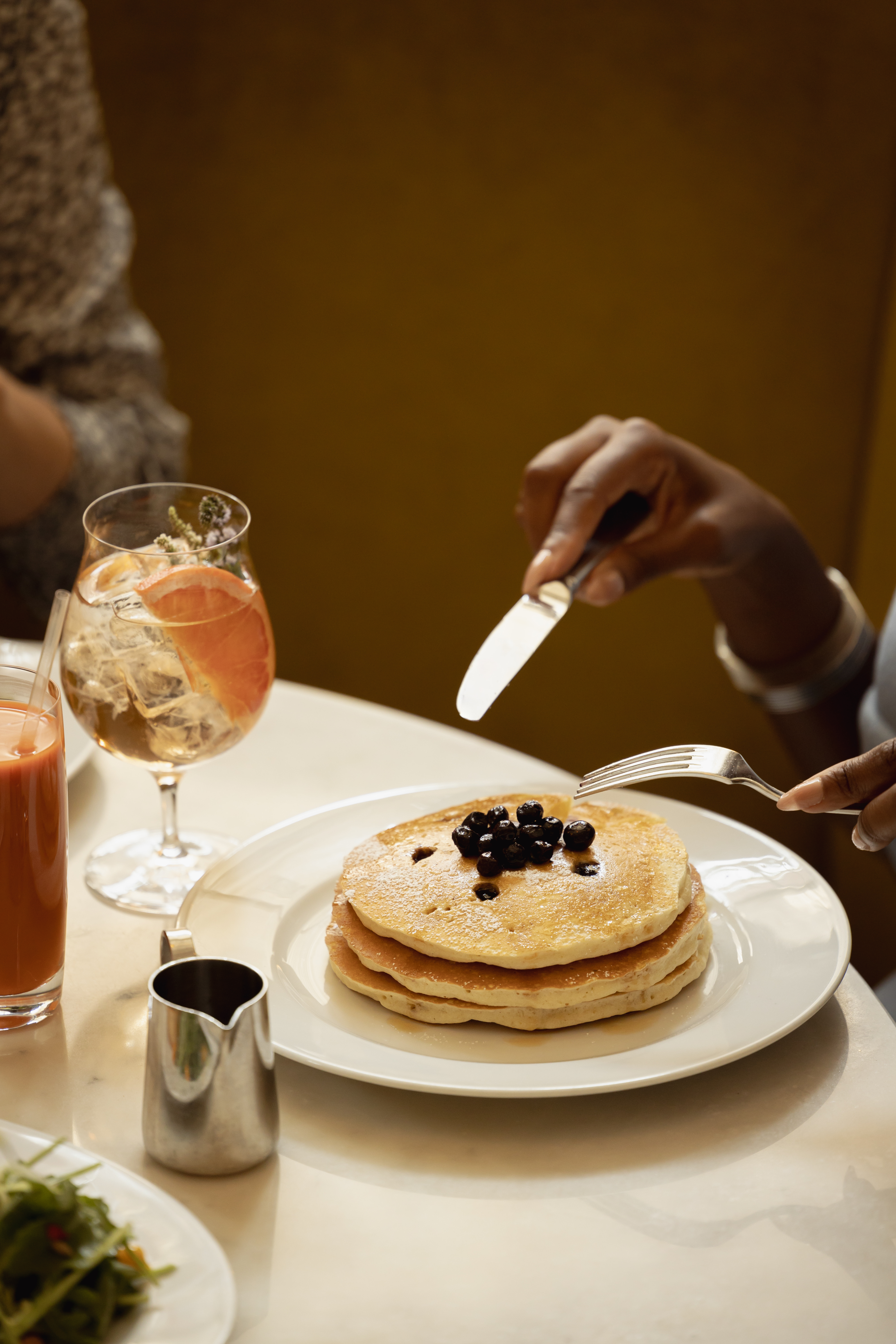 A stack of golden pancakes topped with blueberries is served alongside citrus cocktails and fresh juice.