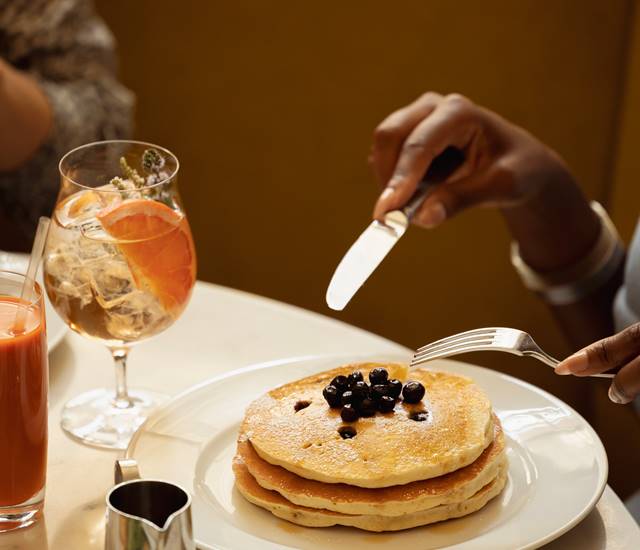 A stack of golden pancakes topped with blueberries is served alongside citrus cocktails and fresh juice.