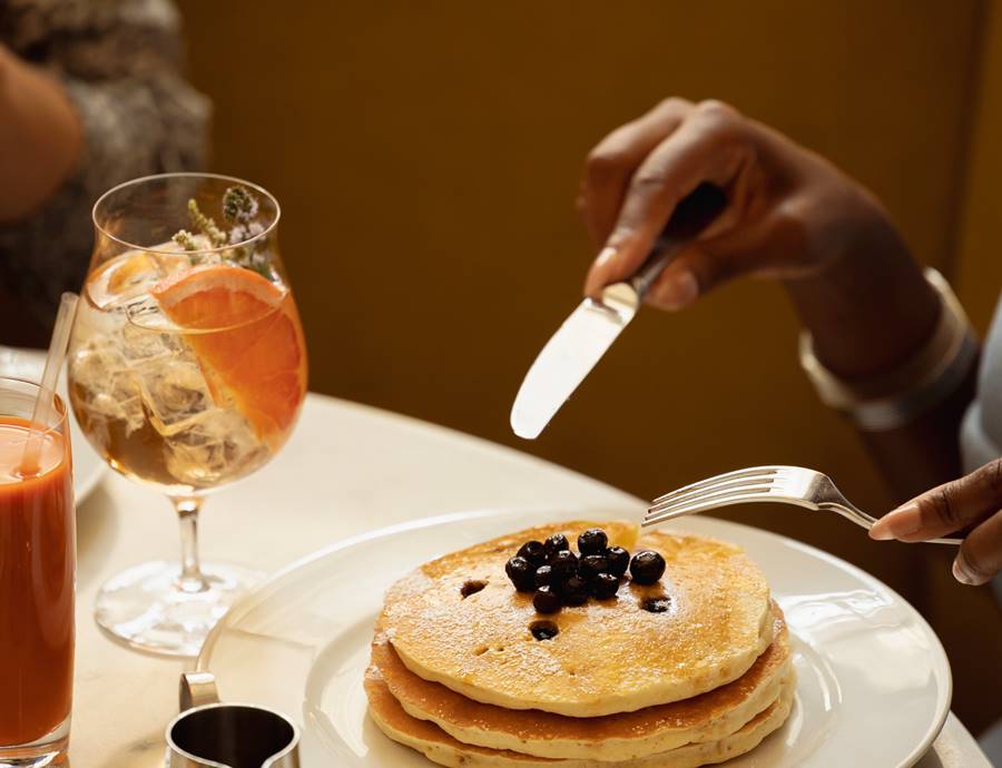 A stack of golden pancakes topped with blueberries is served alongside citrus cocktails and fresh juice.