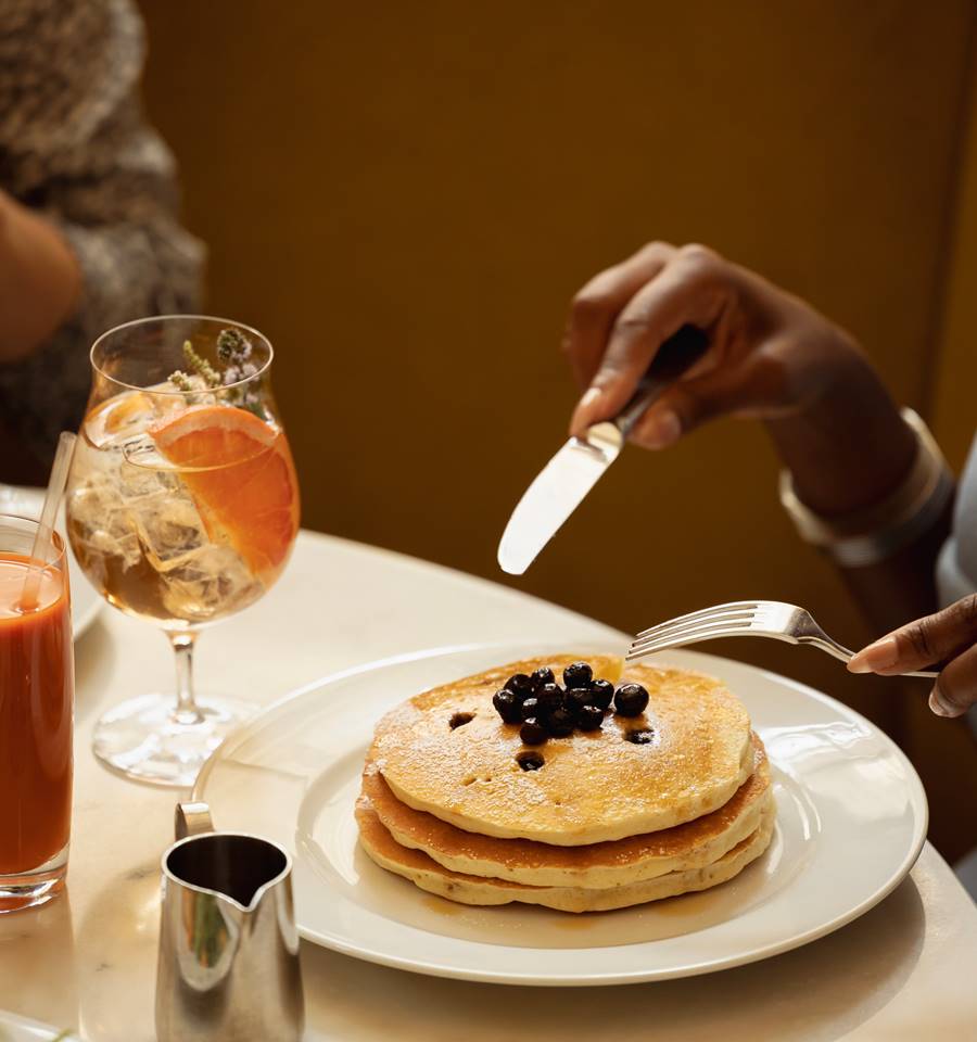 A stack of golden pancakes topped with blueberries is served alongside citrus cocktails and fresh juice.