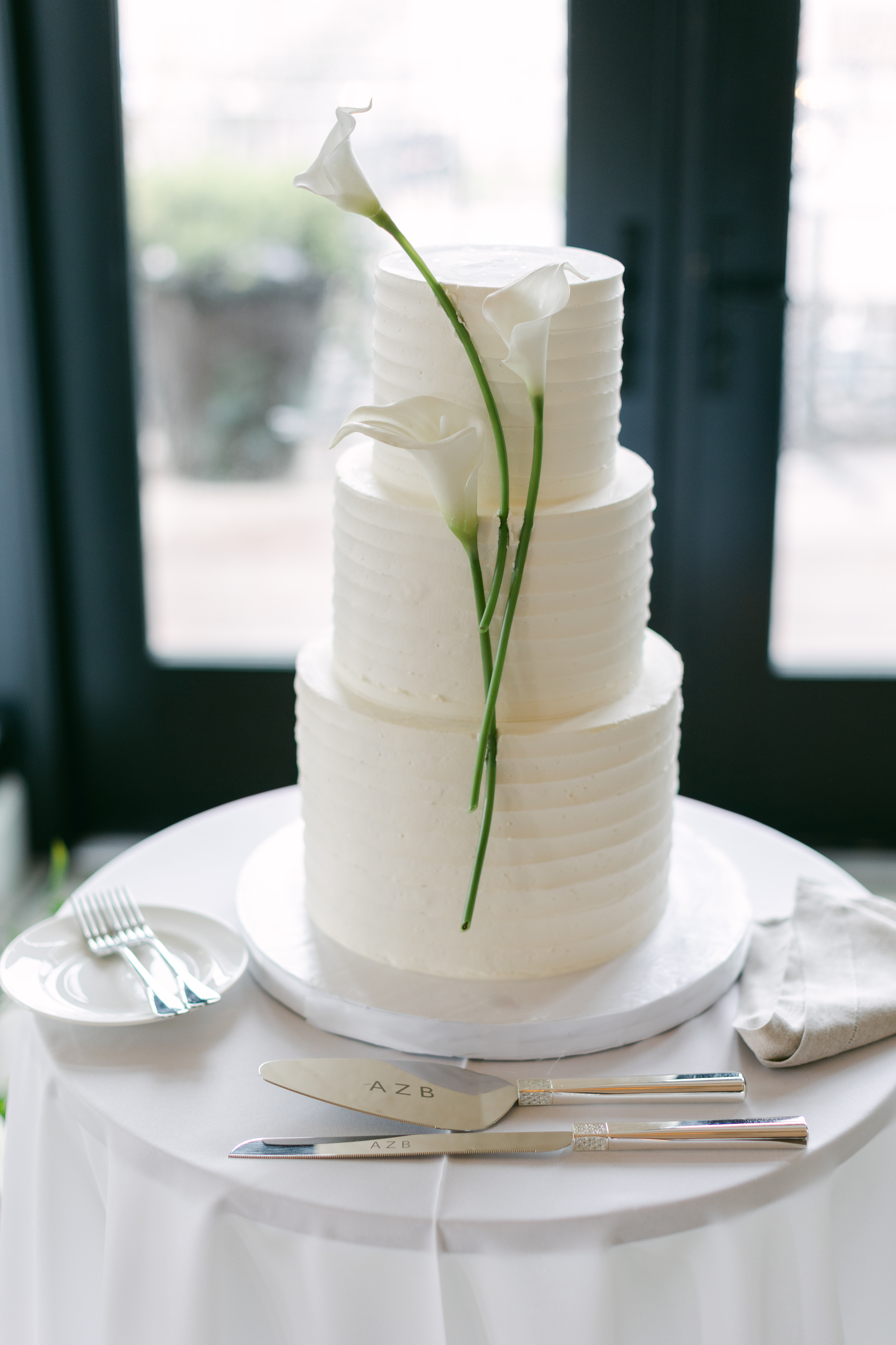 Three-tier white wedding cake with calla lily stems on a round table with silver utensils at a Riggs DC hotel wedding.