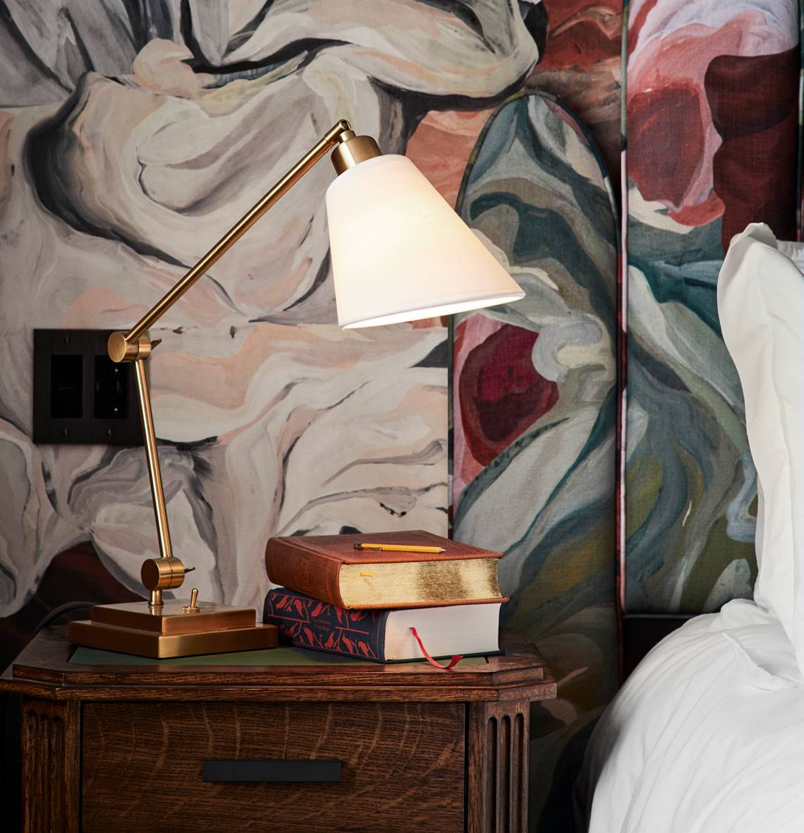 Bedside table at a Riggs DC hotel room with stacked books, a brass reading lamp, and abstract floral wallpaper backdrop.