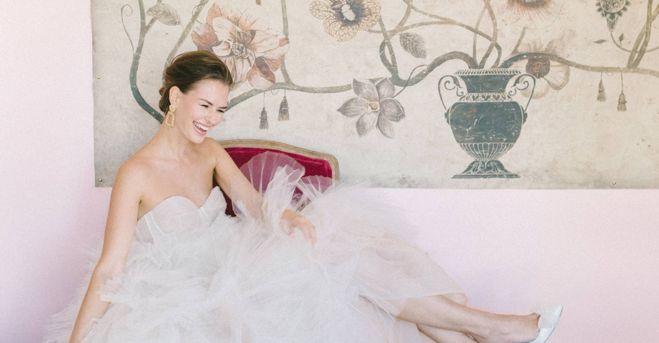 A woman in a white dress sitting on a red chair in front of a pink wall with artwork.