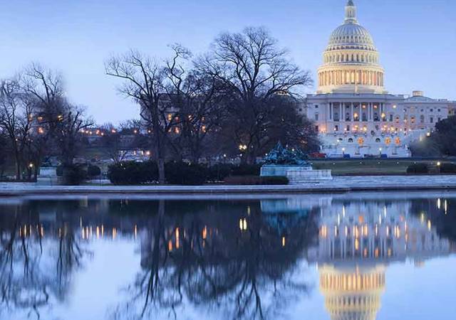 The U.S. Capitol building glows at dusk, its reflection shimmering in the water, framed by leafless trees.