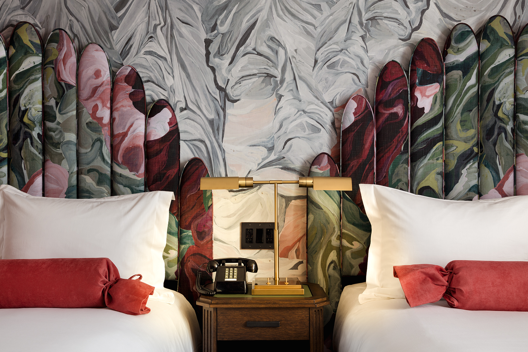 Two hotel beds with floral-patterned headboards, white linens, coral bolsters, and a wooden nightstand with a brass lamp.