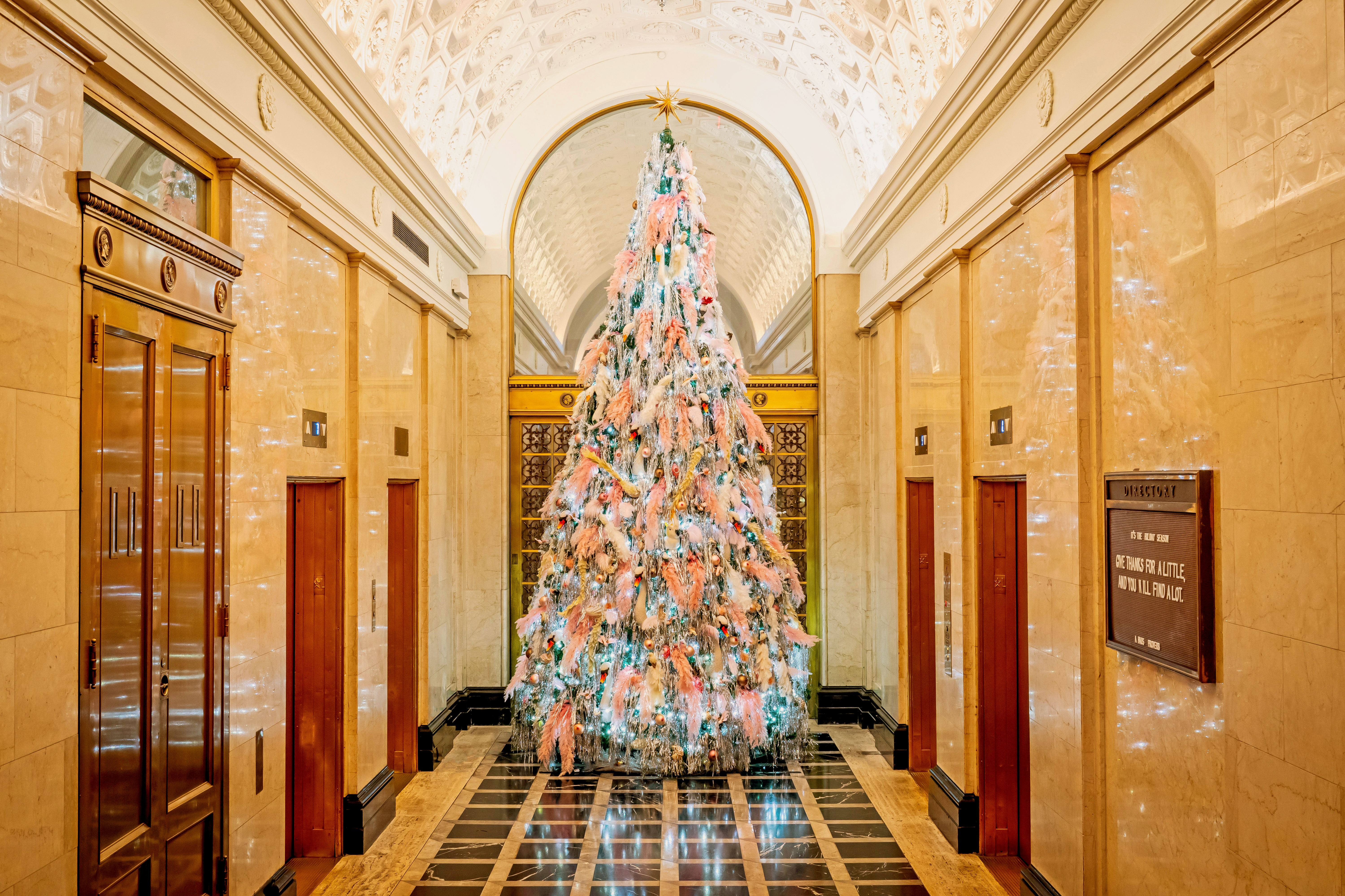 A large Christmas tree stands in a lobby with a tiled floor, arches, and elevators.