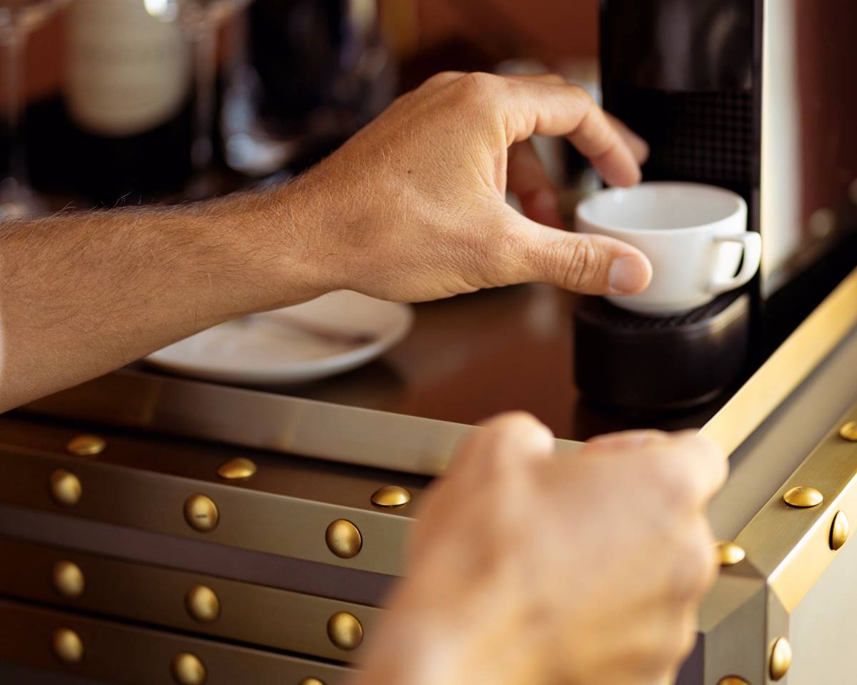 A close-up of a hand placing a small white espresso cup under a coffee machine, with brass-riveted details on the counter.