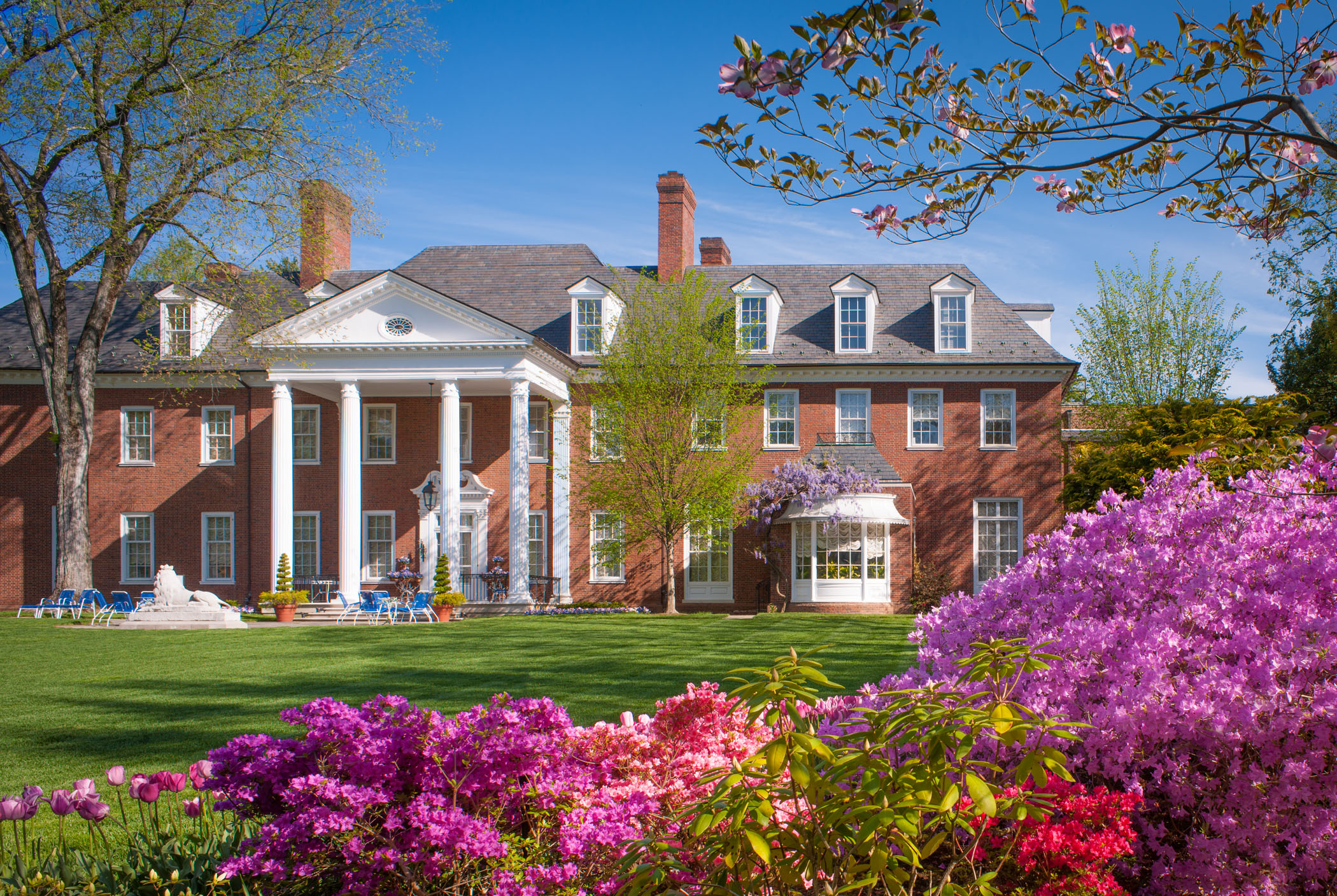 A large brick building with white columns and a gray roof, surrounded by a lawn and flowers.