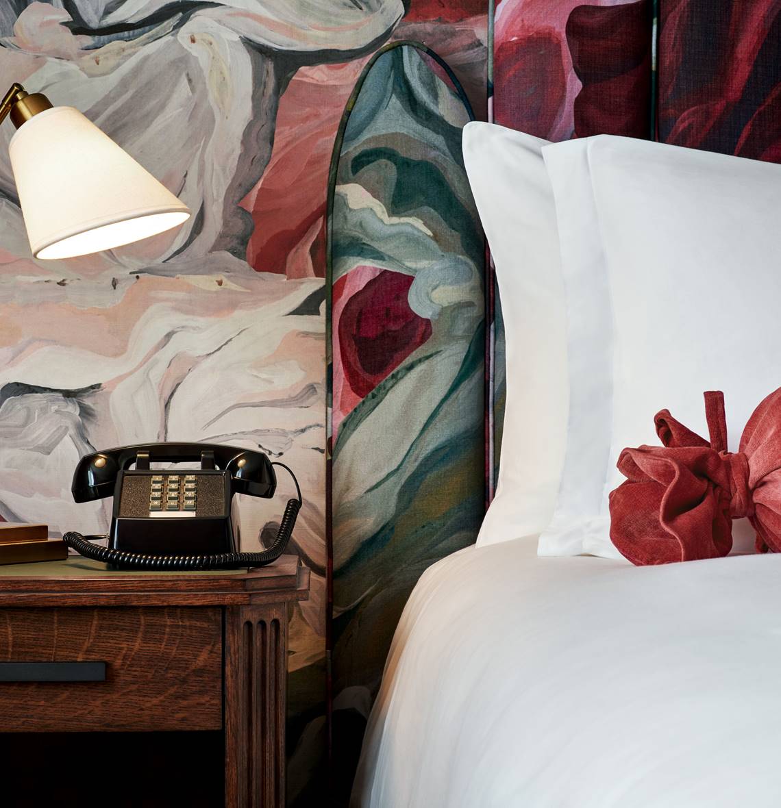 Close-up of a bedside table in a hotel room with a classic black telephone, a brass lamp, and a colorful floral wallpaper.