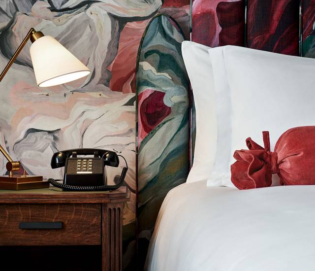 Close-up of a bedside table in a hotel room with a classic black telephone, a brass lamp, and a colorful floral wallpaper.