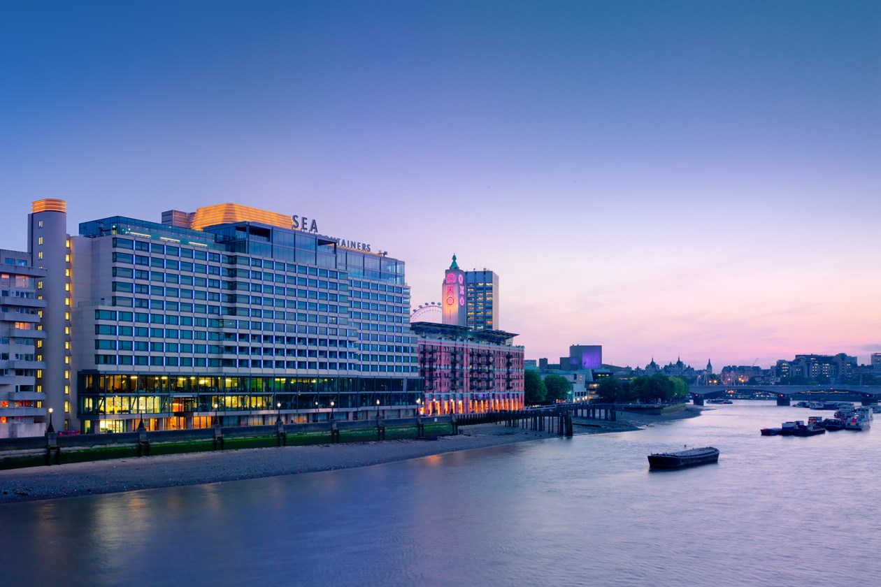 A riverside hotel at dusk with "SEA CONTAINERS" signage, illuminated windows, and the city skyline in the background.