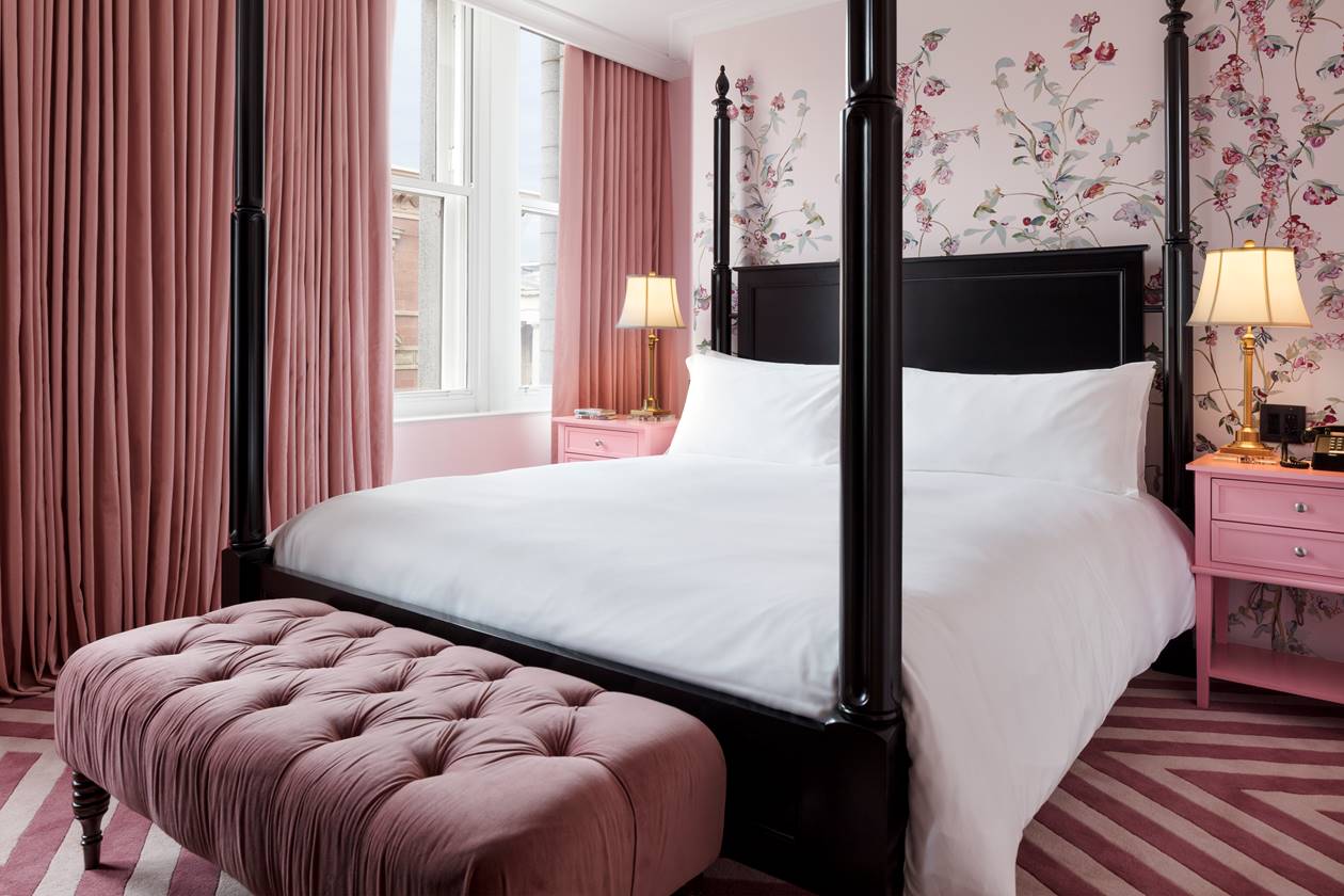 Luxurious bedroom at Riggs DC with a black four-poster bed, pink curtains, a matching pink bench, and floral wallpaper.
