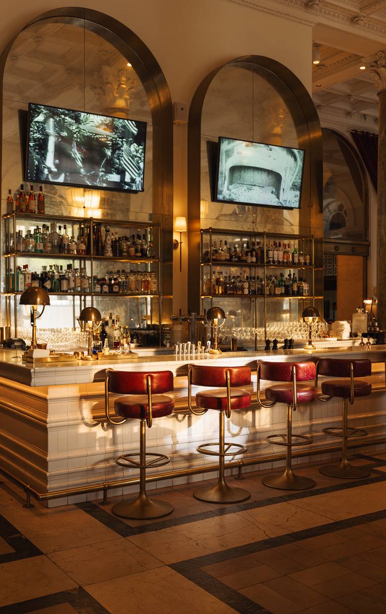 A well-lit bar with a long counter, red stools, and two large TVs on the wall.