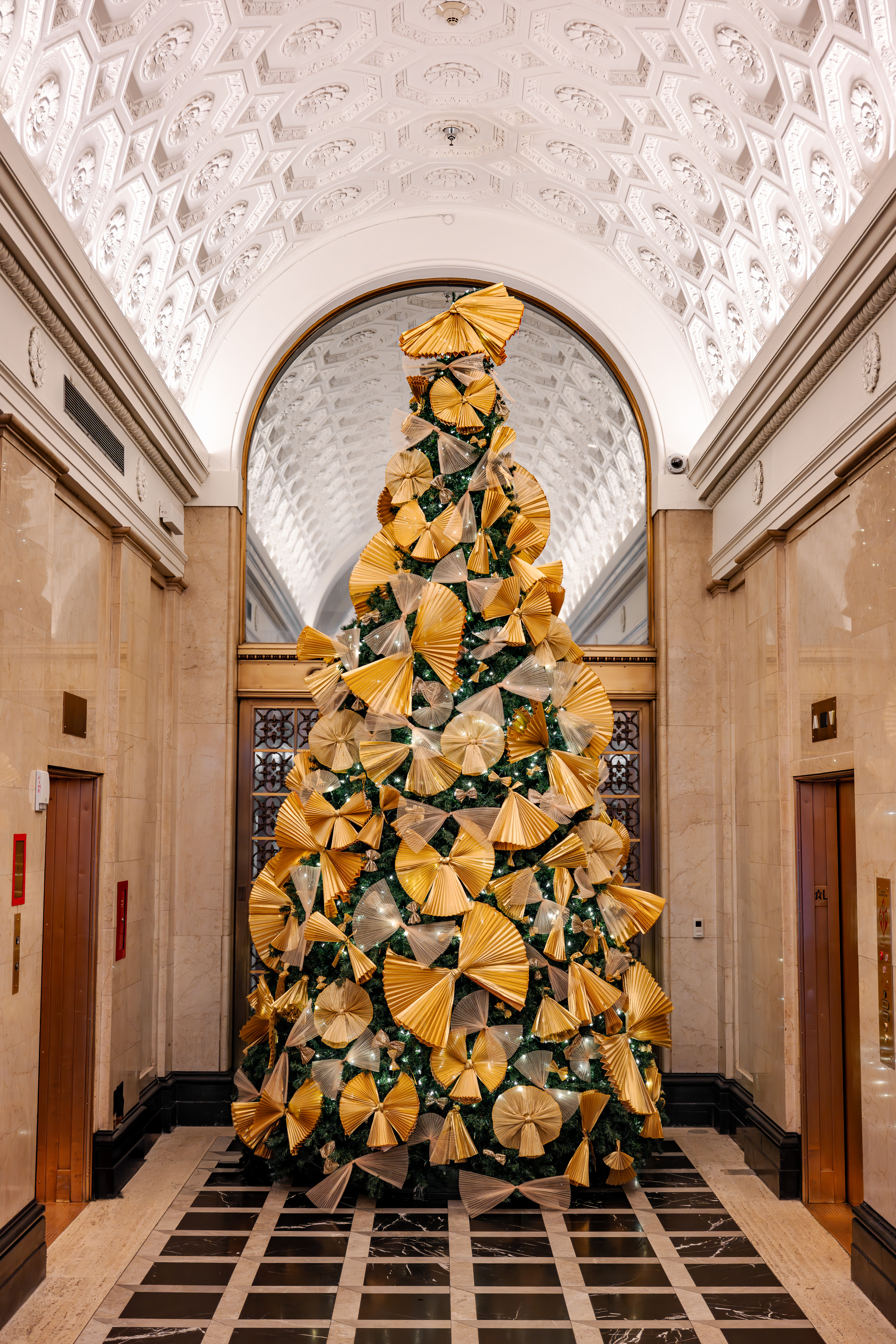 A large Christmas tree decorated with gold and silver ornaments stands in the lobby of a building with high ceilings and marble walls.