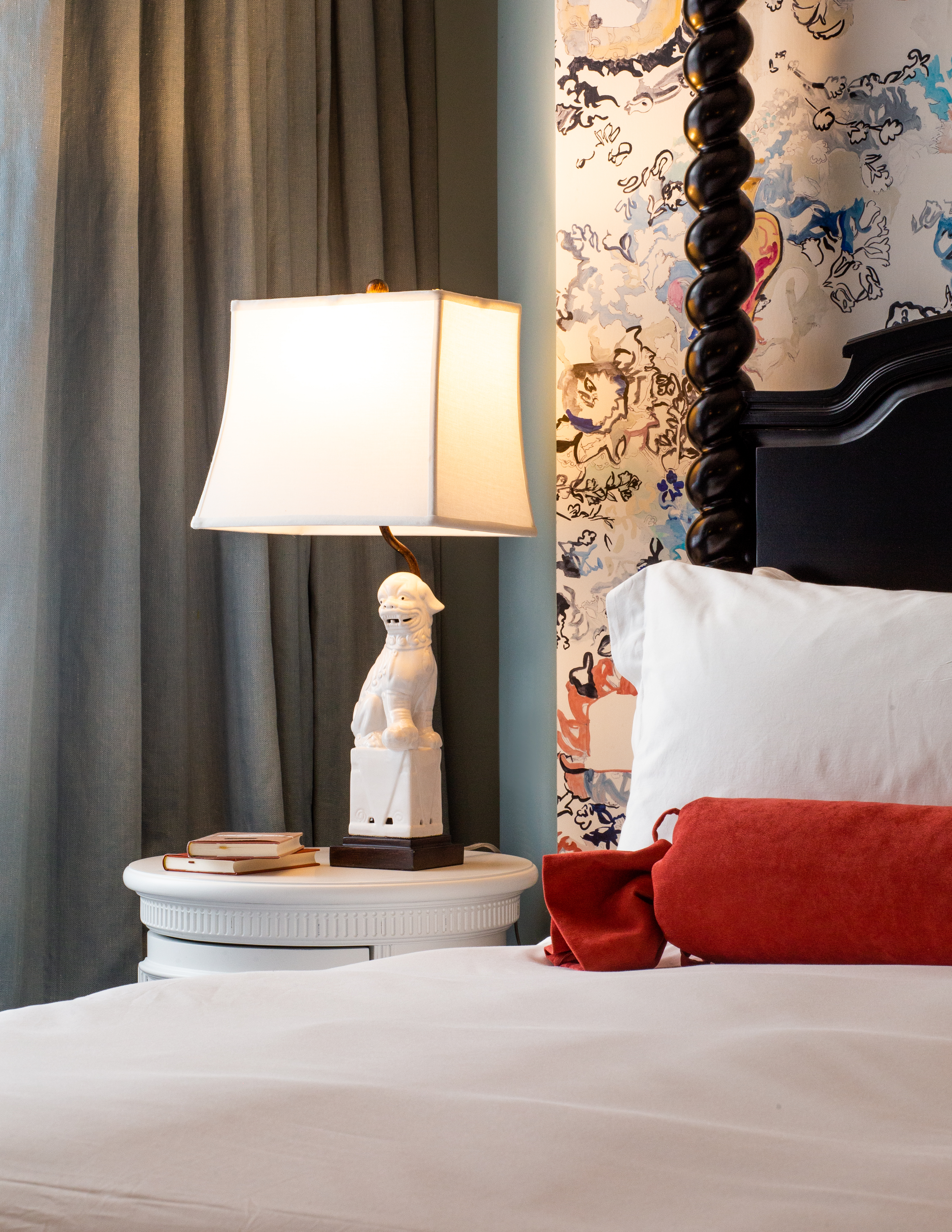 A bedside table with a white lamp featuring a ceramic lion base, against floral wallpaper and blue drapes.