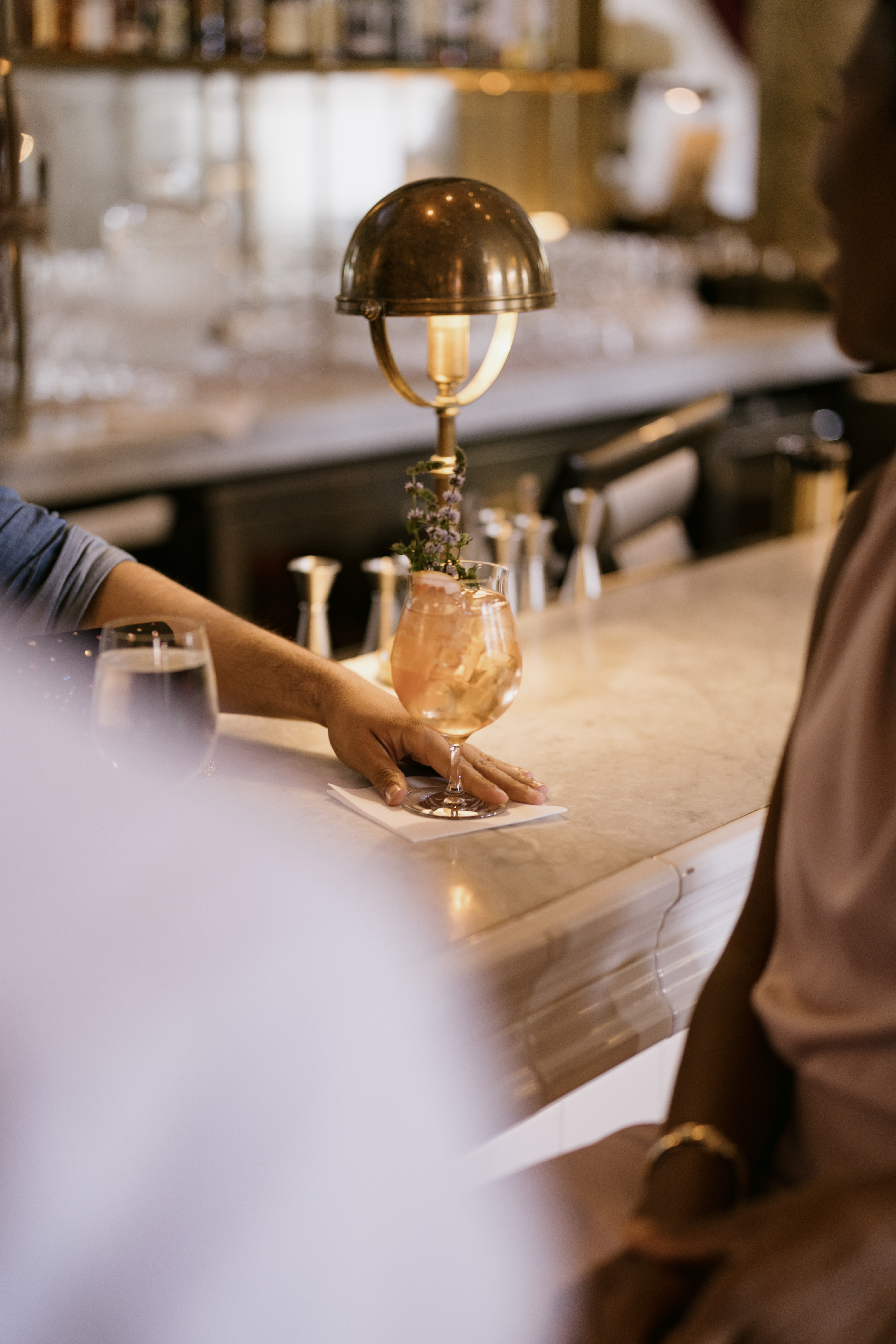 A bartender places a cocktail on the counter, with a brass lamp casting a warm glow in a lively bar setting.