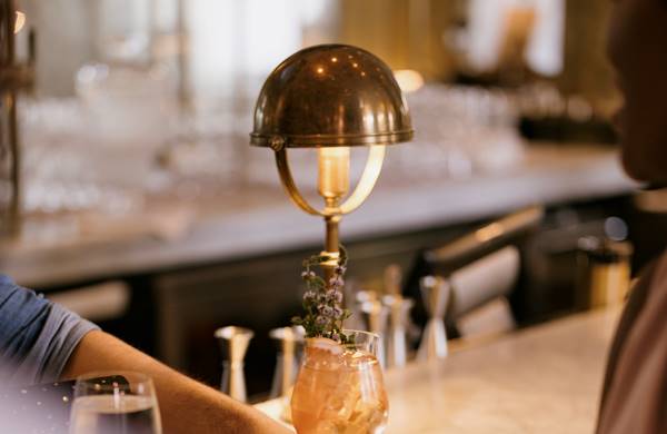 A bartender places a cocktail on the counter, with a brass lamp casting a warm glow in a lively bar setting.