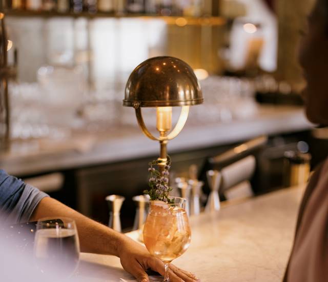 A bartender places a cocktail on the counter, with a brass lamp casting a warm glow in a lively bar setting.