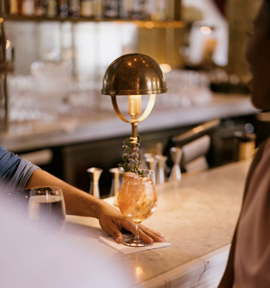 A bartender places a cocktail on the counter, with a brass lamp casting a warm glow in a lively bar setting.