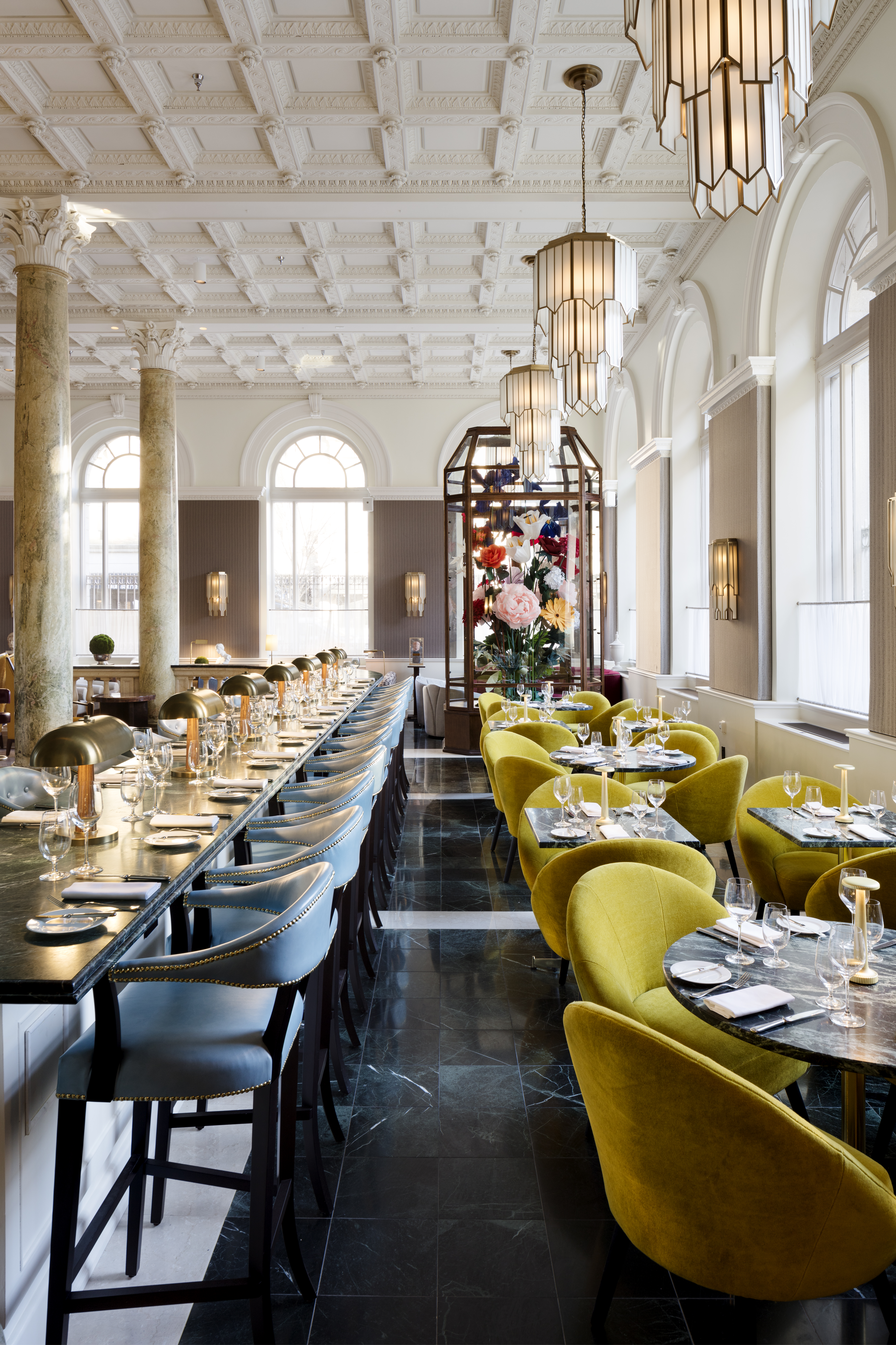 A brightly lit restaurant dining area with long tables set for meals, surrounded by yellow chairs and marble tabletops, with a flower arrangement in the background.