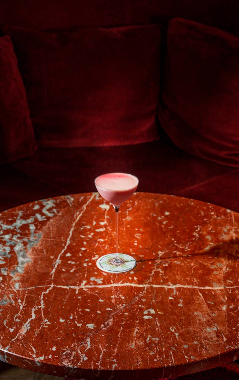 A pink cocktail in a coupe glass sits on a red marble table, against a plush red velvet sofa at the Silver Lyan bar in DC.