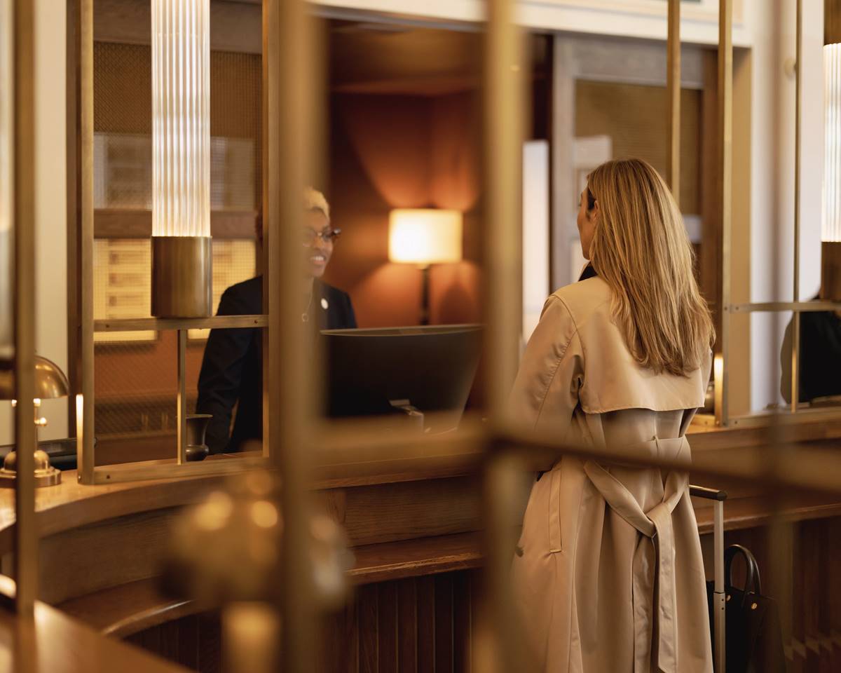 Woman in a trench coat at the hotel front desk of Riggs DC, speaking with a smiling hotel staff member behind a brass-framed counter.